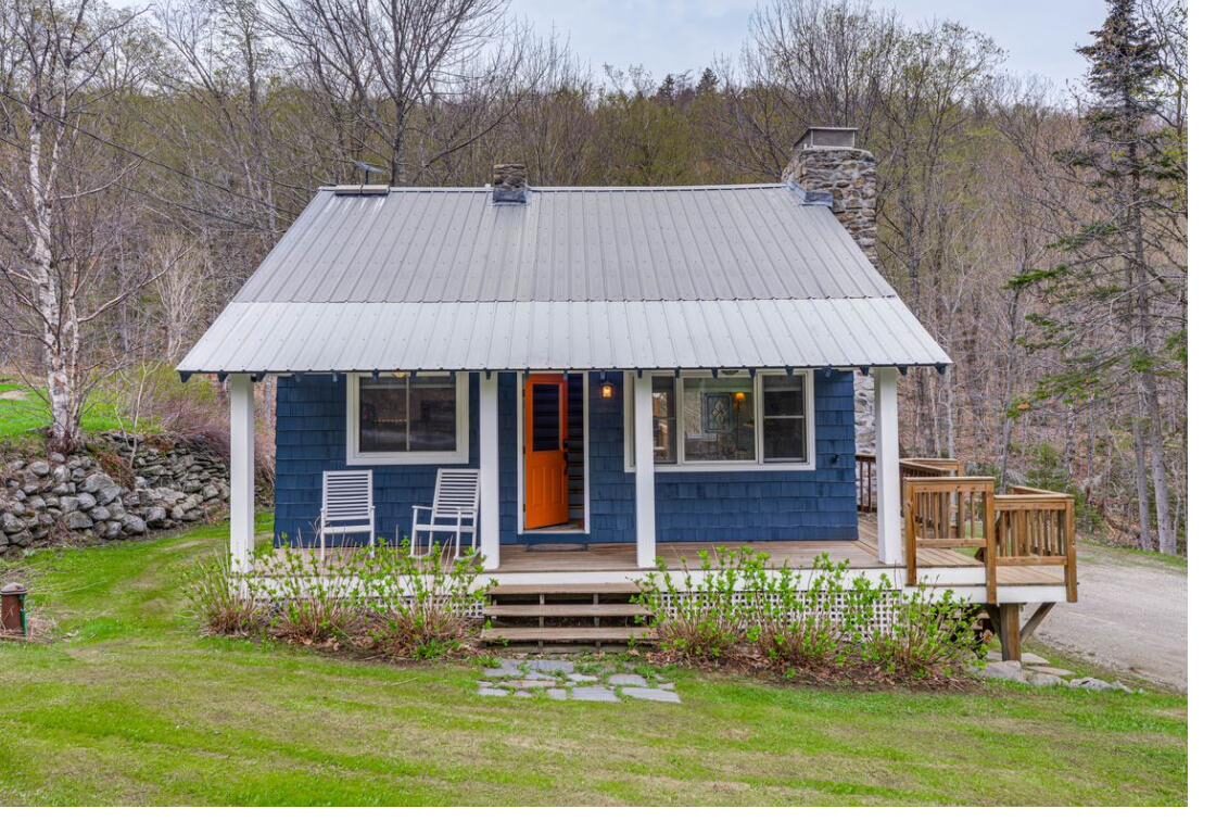 Bluebird Cottage in summer: deep blue siding, a stone chimney, and a front porch surrounded by green Vermont woods